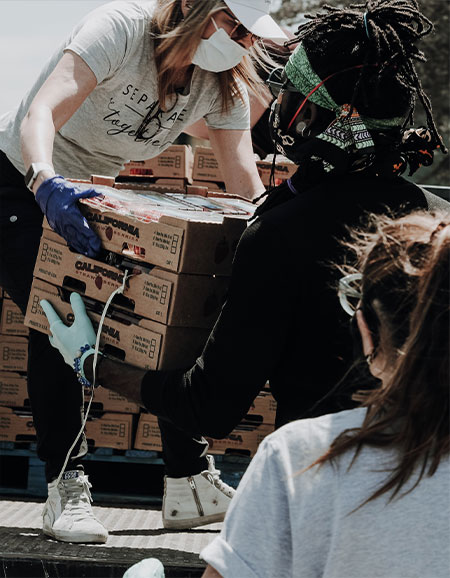 woman in mask handing carton of food to a volunteer while another woman looks on